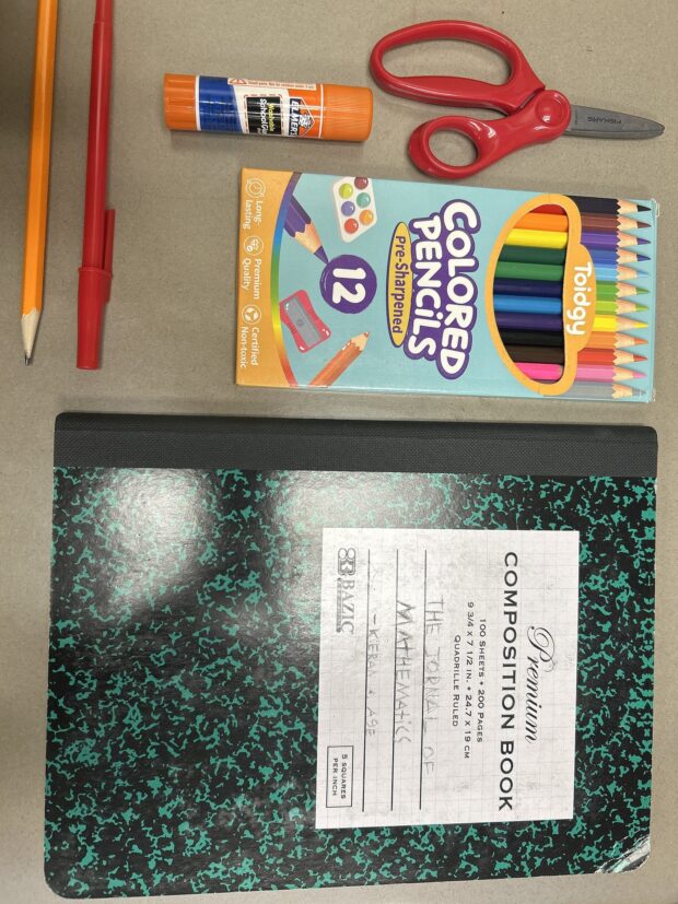 Fournitures scolaires disposées sur une table : crayon, stylo, bâton de colle, ciseaux, boîte de crayons de couleur et cahier.
