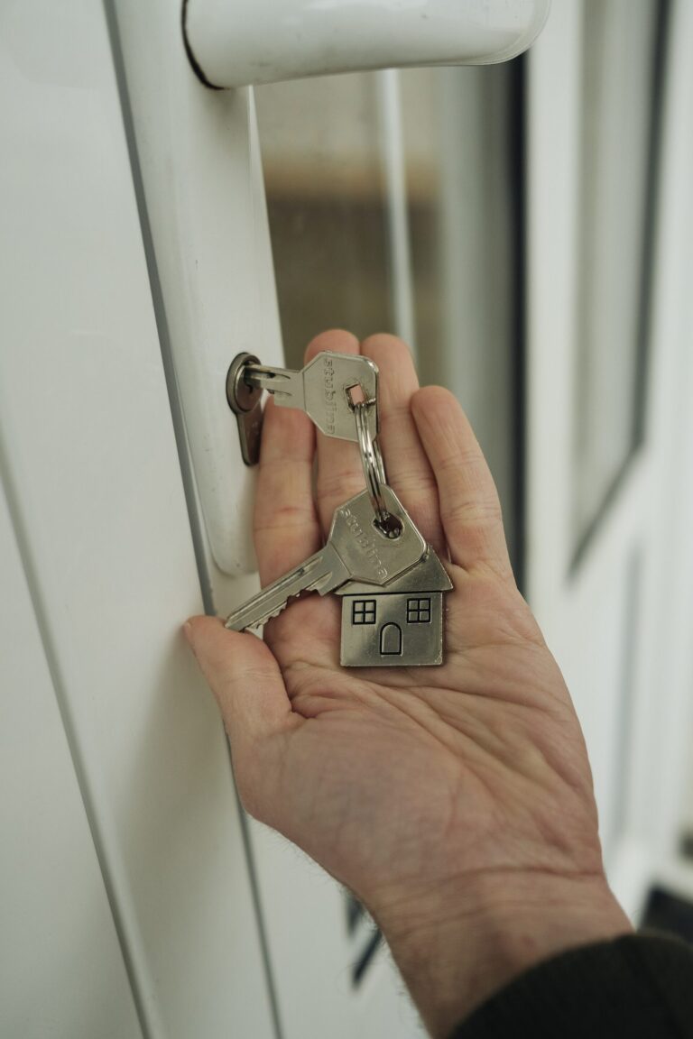 Une main tient un trousseau de cles avec un porte-cles en forme de maison devant la serrure d'une porte