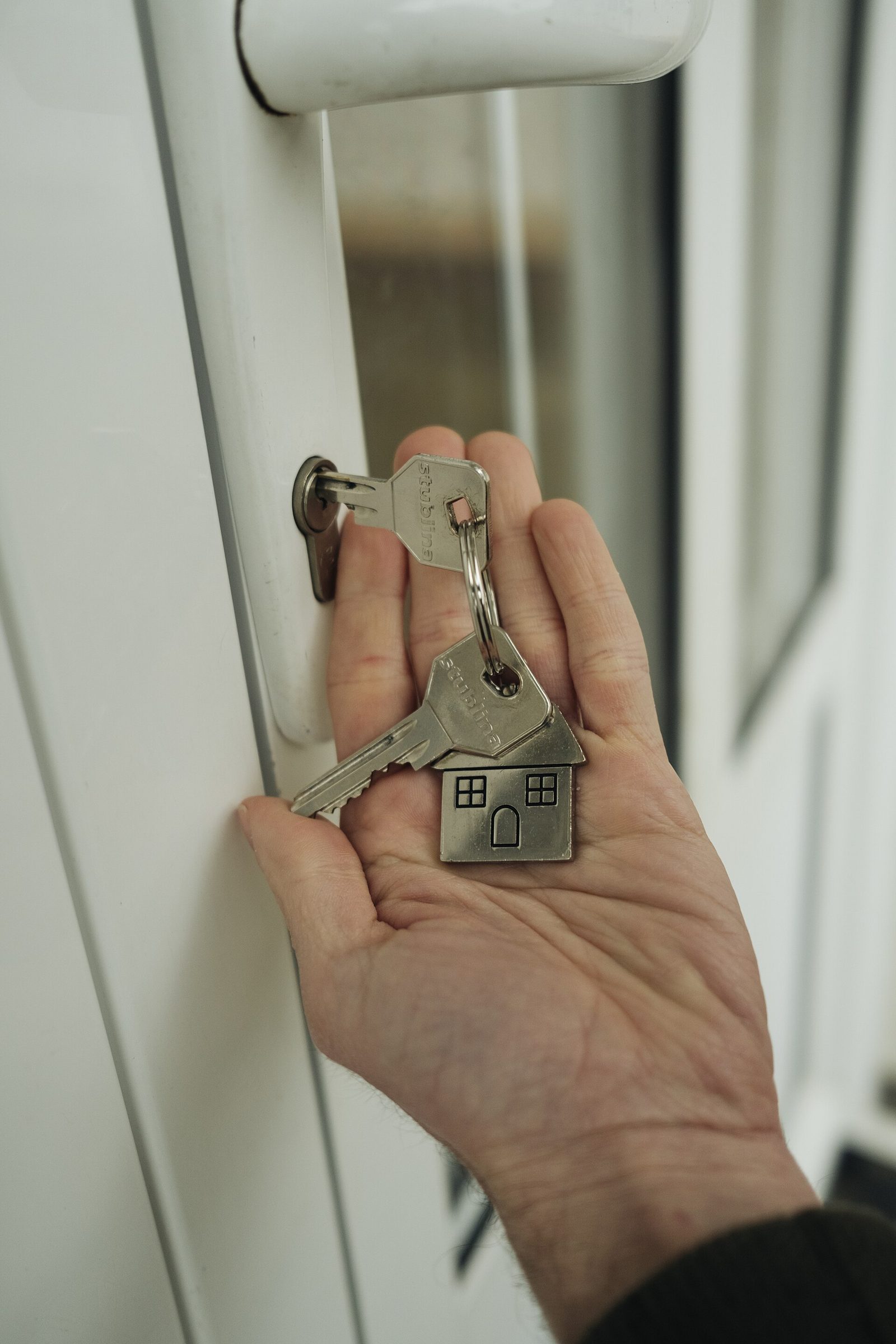 Une main tient un trousseau de cles avec un porte-cles en forme de maison devant la serrure d'une porte