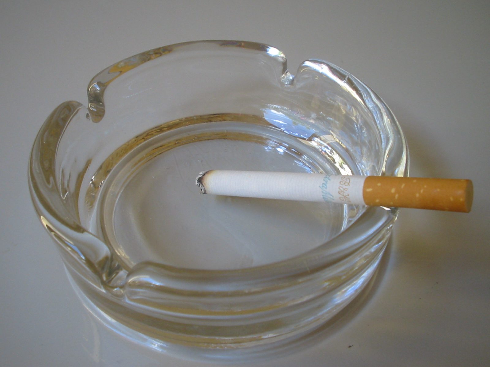 Une cigarette posee sur un cendrier en verre transparent, sur fond clair.