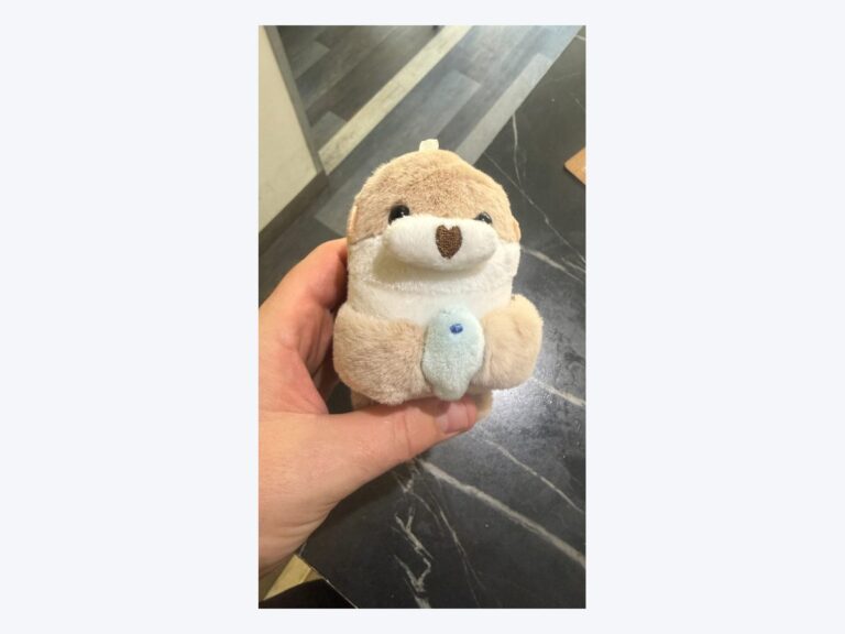Porte-cles en peluche en forme de loutre beige clair tenant un petit poisson bleu, muni d'un mousqueton : le modele concerne par le rappel.